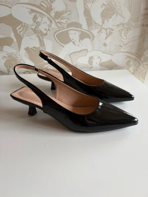 Svarta klackar - Stilrena svarta pumps med lackad finish och spetsig tå. Skorna har öppen häl med smal rem och låg klack, vilket ger en elegant siluett. Är väldigt bekväma och stilrena🩷
