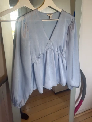 Ljusblå v-ringad blus  - Trendig ljusblå blus från NA-KD med v-ringning och voluminösa långärmar. Blusen har en lös passform med markerad söm under bysten och är tillverkad i ett glansigt, mjukt material som ger en lyxig känsla. Perfekt för att lyfta vilken outfit som helst.