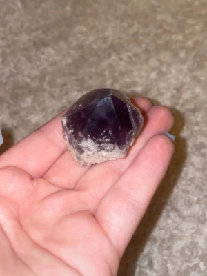 Mörklila rå ametistkristall - Säljer en mörklila ametistkristall med naturlig, rå form och flera tydliga fasetter. Kristallen har en djup lila färg och en ljusare, stenig bas. Perfekt som dekoration, samlarobjekt eller för kristallentusiaster.