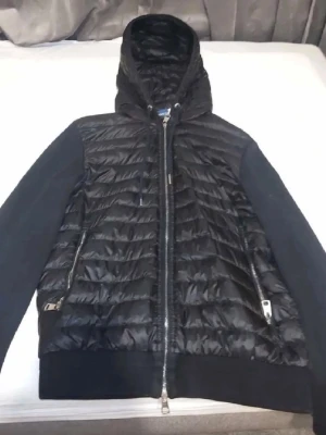Svart Moncler cardiganjacka med huva - Snygg svart cardiganjacka från Moncler med quiltad framsida och huva. Ärmarna är i mjukt tyg medan kroppen är i glansigt, vadderat material. Två dragkedjefickor framtill och ribbade muddar. Perfekt för lager-på-lager och streetwear.