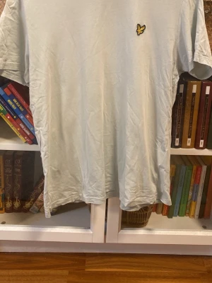 Ljus t-shirt från Lyle & Scott - Säljer en ljus t-shirt från Lyle & Scott med deras klassiska gula fågel-logga broderad på bröstet. T-shirten har korta ärmar och en enkel, clean design som passar till det mesta. Perfekt för dig som gillar stilrena plagg med diskreta detaljer. Finns två små hål på fram och baksidan( se bild 4 och 5)