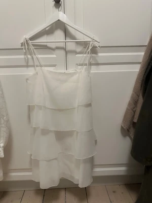 Vit kort klänning med volanger - Säljer en vit kort klänning med tunna axelband och flera lager volanger. Klänningen har en luftig och lätt känsla, perfekt för varma dagar. Axelbanden är knytbara för justerbar passform och modellen är ärmlös.