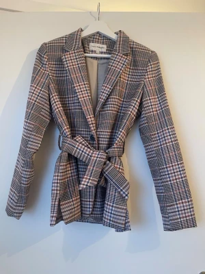 Rutig kavaj med knytskärp KappAhl - Snygg rutig kavaj från KappAhl i beige, svart och rosa toner. Kavajen har klassisk krage, knytskärp i midjan och är helfodrad. Perfekt för dig som vill ha en trendig och stilren look med lite extra edge.