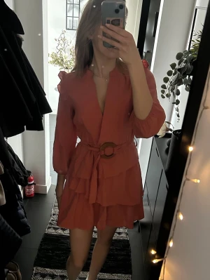 Korall/aprikos omlottklänning med volanger - Trendig kort omlottklänning i korallrosa med volangdetaljer och trekvartsärm. Klänningen har ett bälte med rund spänne i midjan och en lätt v-ringad halsringning. OBS klänningen är med aprikos färgad i verkligheten än vad som framgår på bild🥰