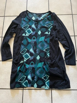 Svart mönstrad långärmad klänning storlek 42/44, bomull  - Cool svart klänning med lång ärm och grafiskt mönster i grönt, blått och turkost framtill. Klänningen har två fickor framtill och är gjord i mjuk bomull. Perfekt för dig som gillar unika prints och bekväma plagg.