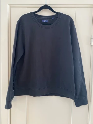 Svart sweatshirt från GANT - Klassisk svart sweatshirt från GANT med rund halsringning och diskret broderad logga på bröstet. Tröjan har långa ärmar och ribbade muddar vid ärmslut och nederkant. Perfekt för en avslappnad och stilren look.Använd och färgen är lite blekare precis vid sömmarna i övrigt i bra begagnat skick  Mått Över bröstet: ca 61 cm Längd från axeln och ner: ca 62 cm  90% bomull och 10% polyester   Kommer från ett djur och rökfritt hem 