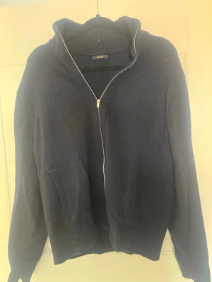 Mörkblå ziptröja från massimoduti - Mörkblå stickad ziptröja  med hög krage och ribbade muddar. Tröjan har två fickor framtill och är tillverkad i ett mjukt, tjockt material som passar perfekt när det är kyligt ute. Enkel och stilren design som funkar till det mesta.