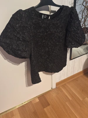 Svart puffärm topp från Pieces - Trendig svart topp från Pieces med puffiga korta ärmar och en unik, skrynklig textur. Toppen har rund halsringning och croppad passform som ger en cool siluett. Använd endast en gång, köpt för ca 499kr