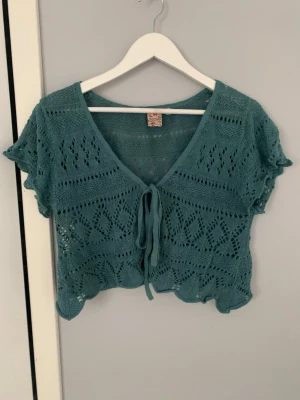 Turkos virkad kortärmad topp - Snygg turkos stickad topp med korta ärmar och knytning framtill. Toppen har ett virkat mönster med hålstickade detaljer och vågig kant nedtill. Perfekt att styla över ett linne eller bikini. Materialet känns lätt och luftigt. Är i storlek medium men funkar för S.
