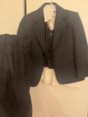 Mörkgrå kostymset med väst och skjorta - Klassiskt mörkgrått kostymset från H&M med kavaj, väst och byxor samt vit skjorta. Kavajen har två knappar och raka slag, västen har tre knappar och byxorna har pressveck. Perfekt för dig som vill ha en stilren och tidlös look.