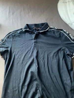 Svart piké från Michael Kors - Snygg svart pikétshirt från Michael Kors med vit detalj på kragen och text längs ärmarna. Klassisk krage, knappar framtill och korta ärmar. Perfekt för en clean och stilren look.