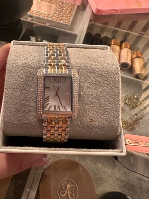 Michael Kors rektangulär klocka guld/silver - Snygg klocka från Michael Kors med rektangulärt ur i silver och guldton. Armbandet är tvåfärgat i silver och guld, och boetten är dekorerad med glittrande stenar runt urtavlan. med plast och lappen fortfarande på. oanvänd men kartongen har några sminkfläckar