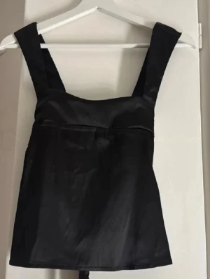 Svart satinlinnetopp med breda band - Snygg svart linnetopp i satin med breda axelband och knyt i ryggen. Toppen har en stilren, minimalistisk look och lätt glansigt material som ger en lyxig känsla. Perfekt att styla med jeans eller kjol för en trendig outfit.