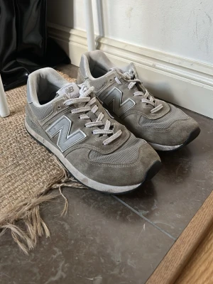 Grå New Balance sneakers i mocka - Snygga grå sneakers från New Balance med klassisk siluett. Skorna har ovandel i mocka och mesh, vita skosnören och den ikoniska N-loggan på sidorna. Perfekta för en avslappnad streetstyle-look. Lite slitna och skosnörena har blivit modifierade.