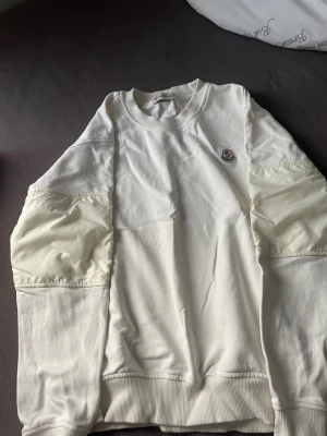 Vit sweatshirt från Moncler - Snygg vit sweatshirt från Moncler med rund halsringning och ribbade muddar. Tröjan har unika detaljer med glansigt tyg på ärmarna och Moncler-logga på bröstet. Perfekt för dig som gillar exklusiva streetwear-plagg. Som ni ser sista bilden en jätte liten skada syns inget när man har på den.