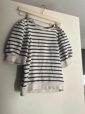 Randig topp från Holly & Whyte XS - Säljer en vit topp med svarta horisontella ränder från Holly & Whyte by Lindex. Modellen har puffiga halvlånga ärmar och breda muddar vid ärmslut och nederkant. Perfekt för en trendig och avslappnad look.