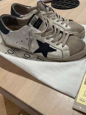 Golden Goose  - Golden Goose sneakers i vitt läder med grå mockadetaljer och marinblå stjärna på sidan. Skorna har beige snören, svart hälparti och GGDB-logga på plösen. Klassisk låg modell med slitna detaljer för en cool streetstyle-look.