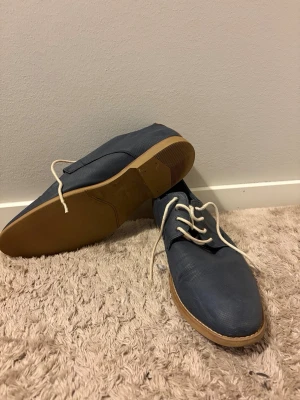 Blå loafers med vita snören - Säljer ett par stilrena blå loafers med vita snören och ljusbruna gummisulor. Skorna har en klassisk form och är tillverkade i ett textilliknande material med diskret struktur. Perfekta för dig som vill ha en clean och avslappnad look.