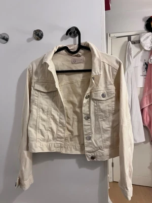 Gul jeansjacka från Lindex - Stilren vit jeansjacka med klassisk krage, två bröstfickor med lock och silverfärgade knappar framtill. Jackan har en rak passform och långa ärmar, perfekt för att lyfta vilken outfit som helst. Finns några fläckar på den, vet tyvärr inte om de går bort i tvätt!