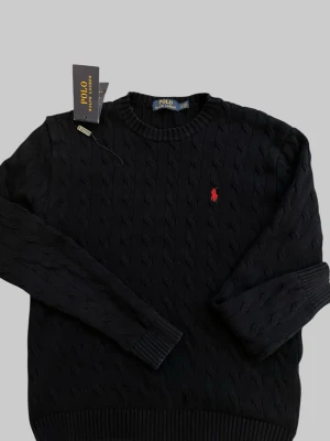  Polo Ralph Lauren tröja  - Kabelstickad svart tröja från Polo Ralph Lauren med rund halsringning och ribbade muddar. Klassisk röd broderad logga på bröstet. Tröjan har en tidlös design och är tillverkad i mjukt material som passar perfekt till många olika stilar jätte fint skick tags medföljer kan gå ner i pris vid snabb affär .