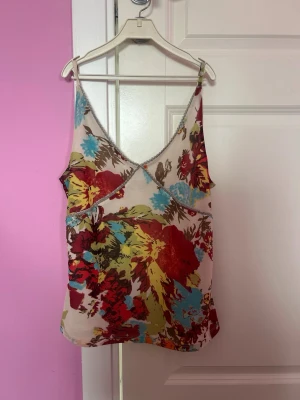 Blommig linnetopp - Säljer en blommig linnetopp med tunna axelband och v-ringning. Toppen har ett färgstarkt mönster i rött, blått, gult och brunt på ljus botten. 