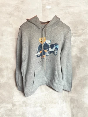 Grå hoodie Polo Bear Ralph Lauren - Säljer en grå hoodie från Ralph Lauren med Polo Bear-tryck framtill där björnen sitter på en bil. Klassisk känguruficka och dragsko i huvan. Mjuk bomullsblandning och avslappnad passform. Perfekt för dig som gillar streetwear och ikoniska prints.
