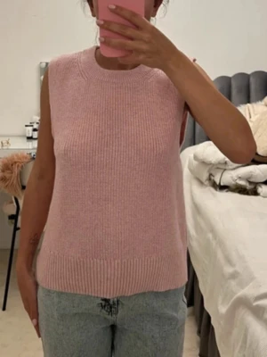 Rosa stickad ärmlös tröja - Säljer en söt rosa stickad ärmlös tröja i mjukt material. Tröjan har rund halsringning och ribbade kanter nedtill och vid halsen. Perfekt att styla över en skjorta eller t-shirt för en trendig lager-på-lager-look.