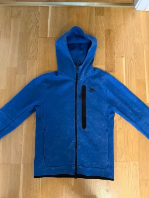 Blå Nike hoodie med dragkedja - Snygg blå hoodie från Nike med hel dragkedja framtill och svart bröstficka med dragkedja. Hoodien har en stor huva och svarta detaljer vid ärmslut och nederkant. Perfekt för en sportig och avslappnad stil. Obs att hoodien ska se urtvättad ut. 