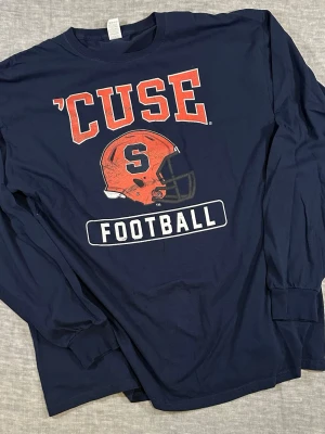 Syracuse ’Cuse football long sleeve shirt XL navy college spell out - Syracuse ’Cuse football long sleeve XL navy. Classic college spell out with orange helmet graphic. 2010s era with a naturally faded print giving it a vintage feel. Relaxed fit, perfect for NCAA / streetwear looks.  Measurements: S2S 50cm, P2P 59cm, Length 76cm, Sleeve 64cm, Waist 58cm.  Used condition with normal wear, print fade adds character.