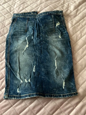 Blå jeanskjol med slitningar - Säljer en blå jeanskjol i klassisk modell med slitna detaljer och rå tvätt. Kjolen har fickor fram och bak samt dragkedja bak. Perfekt för dig som gillar en avslappnad och trendig stil.