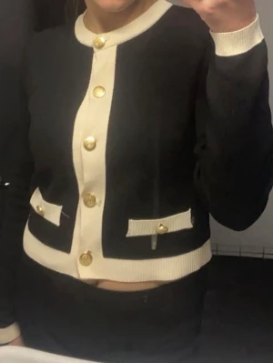 Svart och vit kofta med guldknappar - Stilren svart kofta med vita detaljer och dekorativa guldfärgade knappar framtill och på fickorna. Kortare modell med rund halsringning och ribbade muddar. Perfekt för en chic och trendig look.