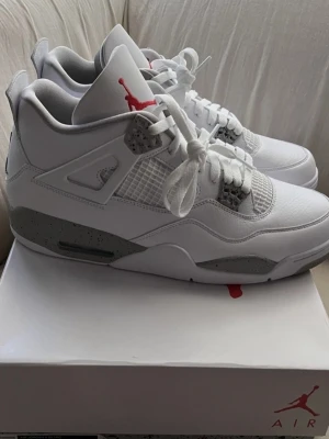 Air Jordan 4 White Cement sneakers - Säljer ett par Air Jordan 4 White Cement sneakers från Nike. Skorna är vita med grå detaljer och svarta stänk på sulan, samt röda inslag på loggan. Klassisk meshpanel på sidan och snörning framtill. Perfekta för dig som vill ha en ikonisk och stilren sneaker.