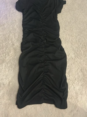 Svart omlottklänning med snörning - Snygg svart klänning med korta ärmar och rynkad snörning längs hela framsidan. Klänningen har en kroppsnära passform och är tillverkad i ett mjukt, stretchigt material som ger en smickrande siluett. Perfekt för dig som gillar stilrena och trendiga plagg.