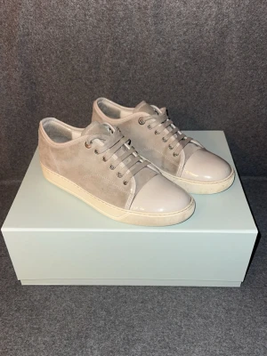 Grå sneakers från Lanvin - Snygga grå sneakers från Lanvin med ovandel i mocka och tåparti i blankt material. Skorna har vita sulor och matchande beige skosnören. Perfekta för dig som gillar stilrena och exklusiva sneakers med en modern touch.