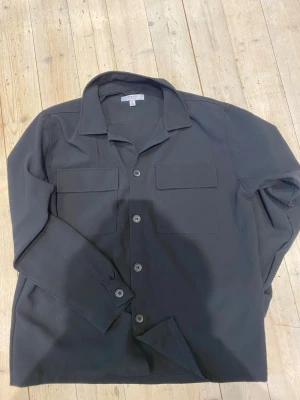 Svart skjorta med bröstfickor - Stilren svart skjorta från COS med klassisk krage och två stora bröstfickor. Skjortan har knappar framtill och långa ärmar, perfekt för en clean och modern look. Materialet känns mjukt och har en lätt struktur.