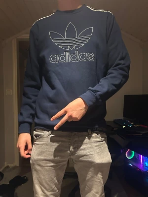 Blå Adidas sweatshirt med logga - Mörkblå sweatshirt från Adidas med stor vit Trefoil-logga och text på bröstet. Tröjan har rund halsringning, långa ärmar och vita detaljer på axlarna. Snygg o passar till allt. Aldrig andvänd men ingen prislapp. PRIS DISKUTERBART