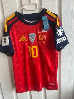 Spaniens landslagströja Lamine Yamal - Röd och marinblå fotbollströja från Adidas, officiell Spanien landslagströja med nummer 10 och Lamine Yamal på ryggen. Tröjan har gula detaljer, tryckta märken och patchar på ärmarna. Perfekt för fotbollsfans som vill sticka ut på matchen.