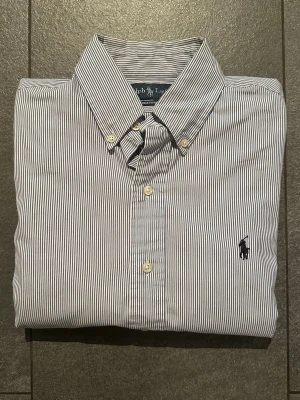Randig skjorta från Ralph Lauren - Klassisk skjorta från Ralph Lauren med smala blå och vita ränder, broderad logga på bröstet och knappar framtill. Skjortan har lång ärm och button-down krage. Perfekt för en stilren look. Skjortan är i gott skick men har en mindre fläck på axeln (se bild)