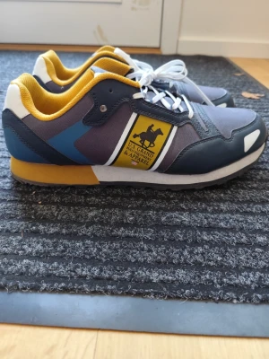 Färgglada sneakers från U.S. Grand Polo - Snygga sneakers i blått, grått och vitt med gula detaljer och vit sula. Märket U.S. Grand Polo syns tydligt på sidan. Skorna har mesh och syntetmaterial, vita snören och en sportig vibe. Perfekta för dig som gillar färg och stil.