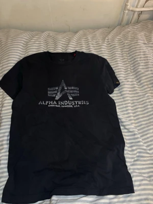 Svart Alpha Industries t-shirt - Snygg svart t-shirt från Alpha Industries med deras ikoniska logga och text framtill. Klassisk rund hals och korta ärmar. Tillverkad i mjuk bomull som känns skön mot huden. Perfekt för dig som gillar streetwear och vill ha en enkel men stilren look.