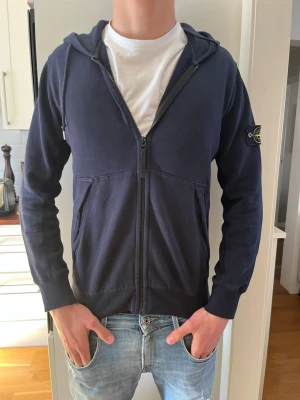 Mörkblå hoodie från Stone Island - Snygg mörkblå hoodie från Stone Island med dragkedja framtill och klassisk logotyp-patch på vänster ärm. Hoodien har huva, två fickor fram och ribbade muddar. Perfekt för en avslappnad och trendig stil. Storlek M men passar S