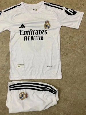 Real Madrid matchställ Mbappé 9 - Snyggt vitt matchställ från Real Madrid med Mbappé och nummer 9 på ryggen. Setet består av tröja och shorts med svarta detaljer och klubbmärke. Adidas-logga och Emirates Fly Better-tryck på framsidan. Perfekt för fotbollsträning eller att bära på läktaren.