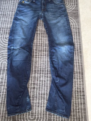 Mörkblå gstar jeans med snygga detaljer - Säljer ett par mörkblå jeans med coola sömmar och slitningar på benen. Jeansen har klassisk femficksdesign och markerade bakfickor med dekorativa detaljer. Perfekta för dig som gillar en avslappnad men trendig stil.