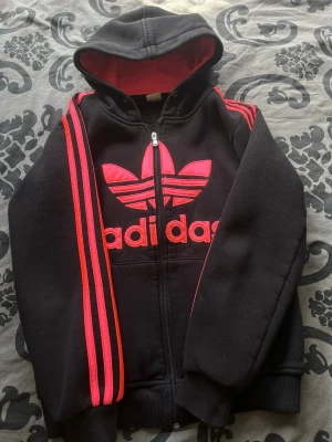 Svart Adidas hoodie med röda detaljer - Svart hoodie från Adidas med dragkedja, stor röd Adidas-logga på bröstet och röda ränder längs ärmarna. Insidan av huvan är röd och materialet är mjukt och skönt, perfekt för en sportig och avslappnad stil.