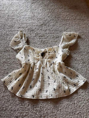 Blommig vit blus med snörning - Söt blus med små blommiga mönster och snörning framtill. Blusen har korta, volangiga ärmar och är tillverkad i ett krinklat bomullsmaterial som känns lätt och luftigt. Perfekt för dig som gillar trendiga toppar.
