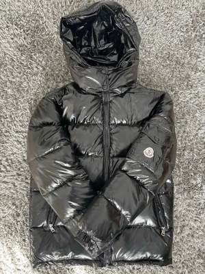 Moncler Maya - Snygg svart dunjacka från Moncler med glansig finish och huva. Jackan har dragkedja framtill, två fickor med dragkedja och en ärmficka med Moncler-logga. Perfekt för kalla vinterdagar och riktigt trendig look.
