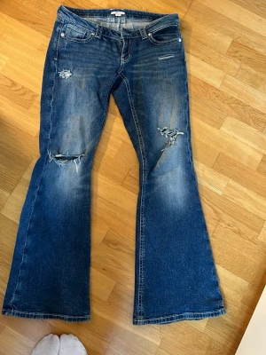 Blå bootcut jeans med slitningar - Säljer ett par blå bootcut jeans från Gina Young 14 + med coola slitningar och distressed detaljer på benen. Jeansen har klassisk femficksdesign och är tillverkade i denim med lite stretch. Perfekt för dig som gillar en avslappnad och trendig look.