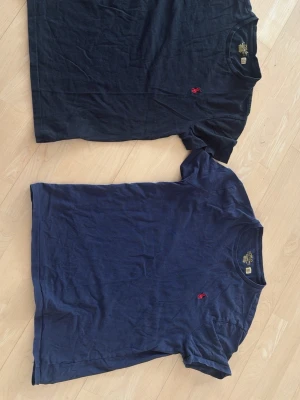 Två t-shirts från Ralph Lauren - Säljer två klassiska t-shirts från Ralph Lauren, en svart och en marinblå. Båda har rund hals och den ikoniska röda logotypen broderad på bröstet. Tillverkade i mjuk bomull som är skön att bära. Perfekta till jeans eller shorts för en enkel och stilren look.