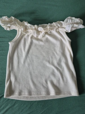 Vit offshoulder topp med volang - Superfin vit offshoulder topp med volang runt axlarna. Toppen är ärmlös och har en lätt ribbad struktur i materialet. Perfekt för dig som vill ha en söt och trendig look till sommaren.