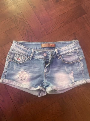 Blå denimshorts med slitningar - Det står att de är i storlek XL men är väldigt små i storlek så skulle säga mer S, kanske M! Säljer ett par blå denimshorts från Dsquared2 med slitningar och coola detaljer som en Union Jack-patch på bakfickan. Shortsens midja är låg och de har klassiska fem fickor samt uppvikta benslut. Perfekta för sommarens alla äventyr.